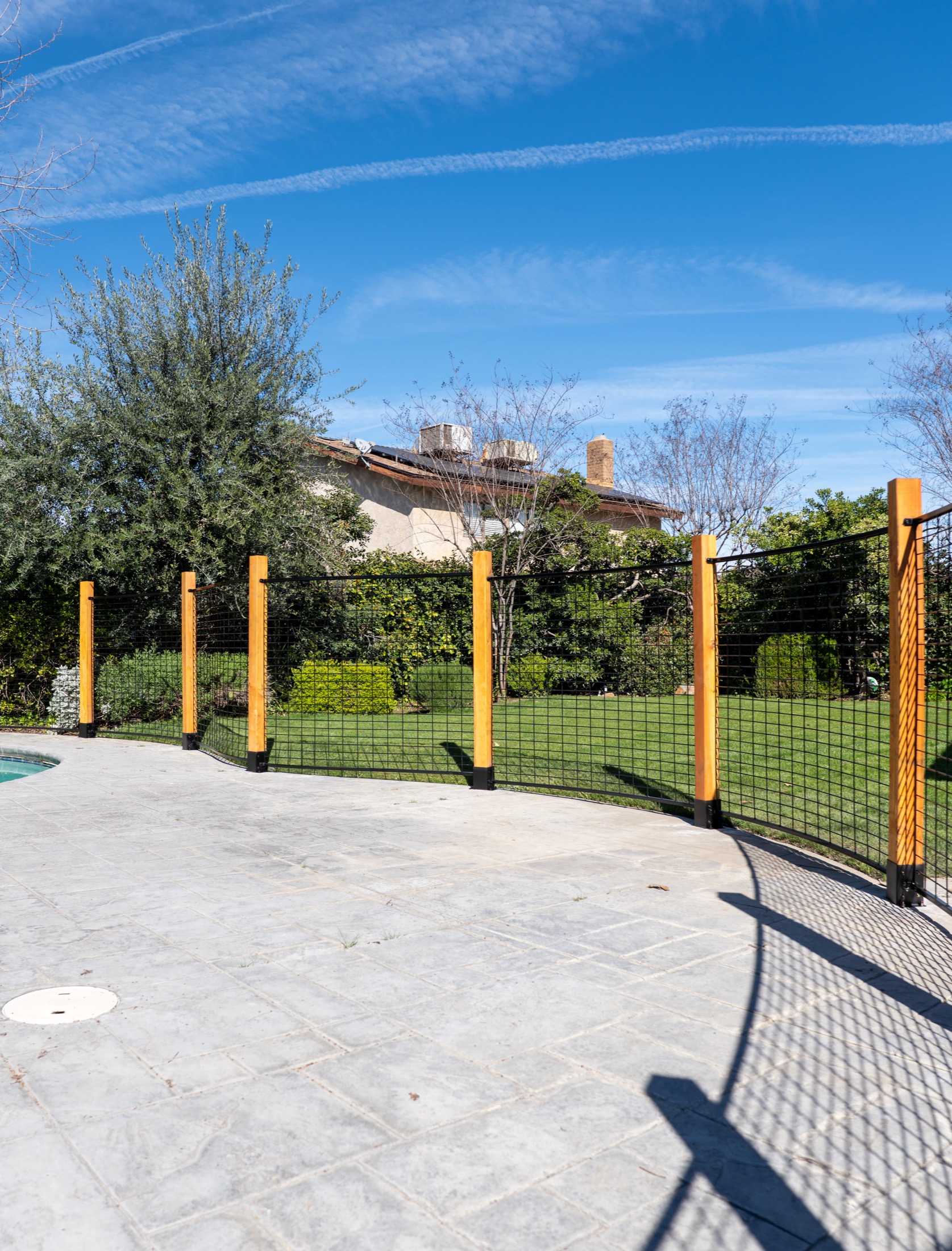 Curved black hog panel pool fence showing wood post spacing, patio edge transitions, and open-view welded wire panels