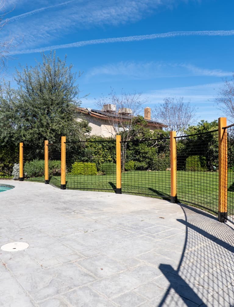 Curved black hog panel pool fence showing wood post spacing, patio edge transitions, and open-view welded wire panels
